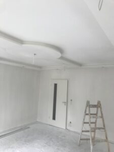 Renovierter Raum mit weißer Tür, Schutzfolie auf Boden, Leiter vor Wand und dekorativem Stuckelement an weißer Decke