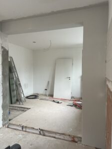 Renovierter Raum mit Leiter, freiliegenden Kabeln, Bohrmaschine, Tür, offenen Bodenbereichen und Wandreparaturen