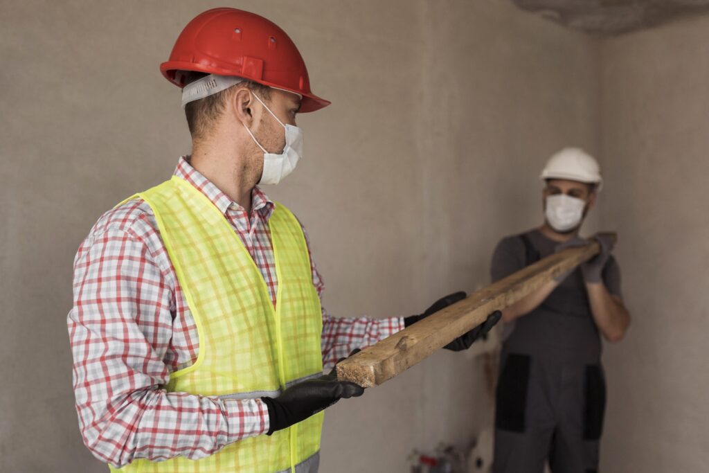 Zwei Bauarbeiter mit Schutzhelmen und Handschuhen heben gemeinsam Holzbohle in Innenraum.