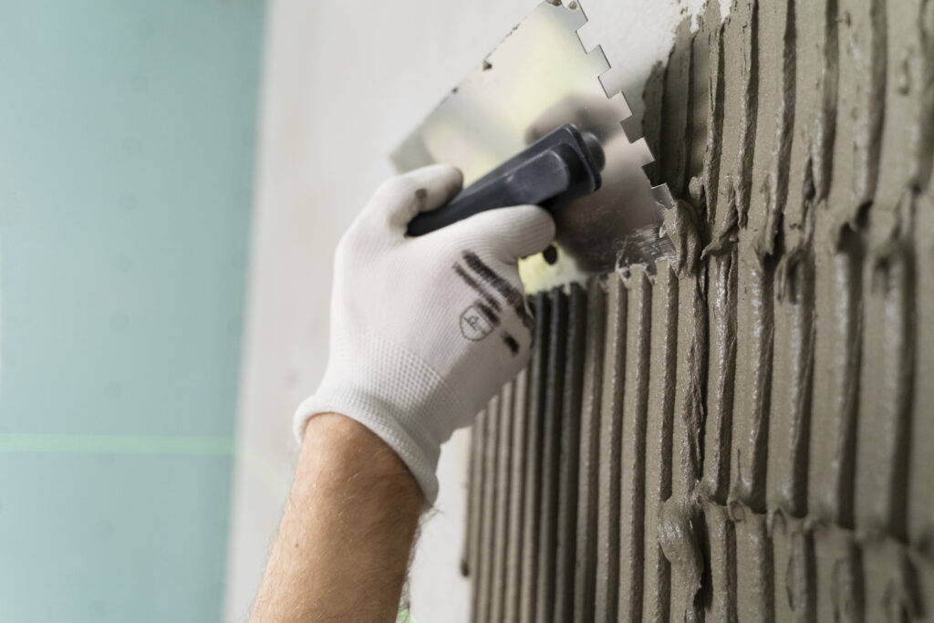 Ein Handwerker trägt mit einer Zahnkelle gleichmäßig Fliesenkleber auf eine Wandfläche auf, er trägt weiße Arbeitshandschuhe.