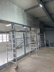 Renovierter Innenraum mit zwei fahrbaren Gerüsten, teils gespachtelten Trockenbauwänden, offener Decke mit Metallträgern und Treppenabgang